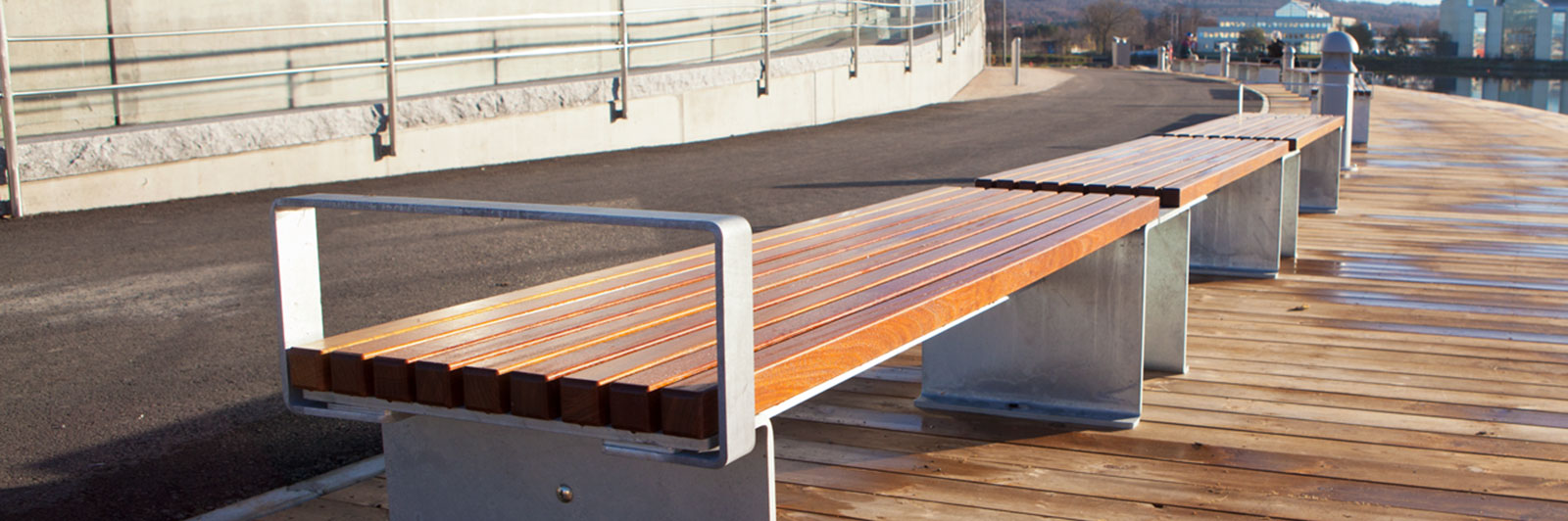 Row of wooden benches with steel armrests and frame, along a walkway by a lake.